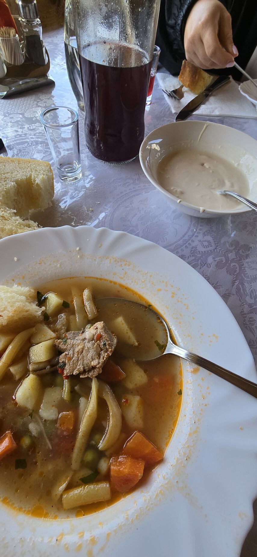 O supă caldă cu legume și carne, servită pe o masă cu băuturi și pâine.