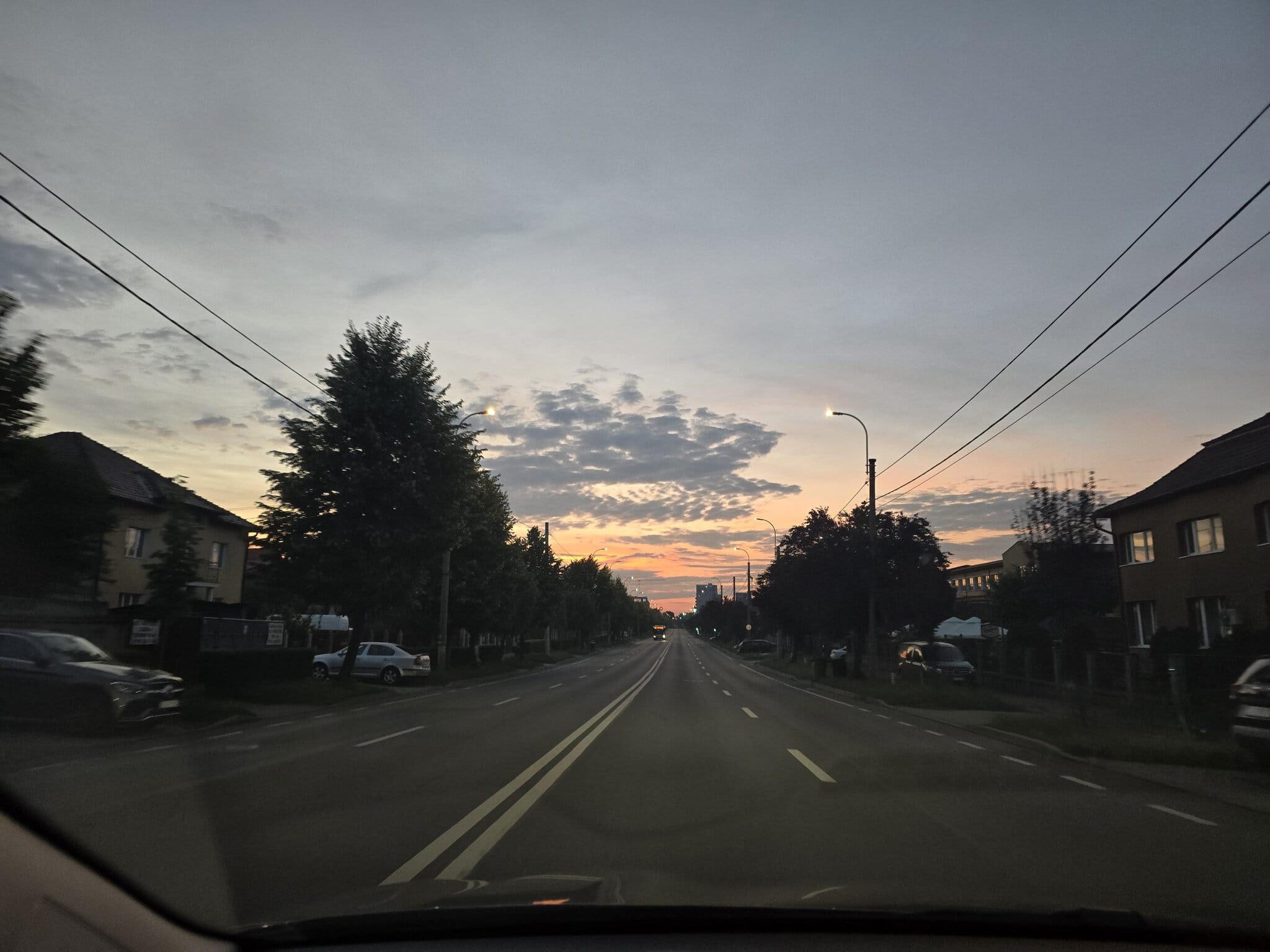 Vedere de pe bord spre o stradă largă de oraș cu apus de soare portocaliu și nori dramatici pe cer.