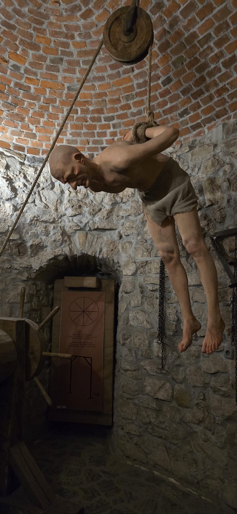 Sculptură realistă a unui prizonier suspendat cu mâinile legate la spate, expusă într-un muzeu medieval cu ziduri de piatră și cărămidă.