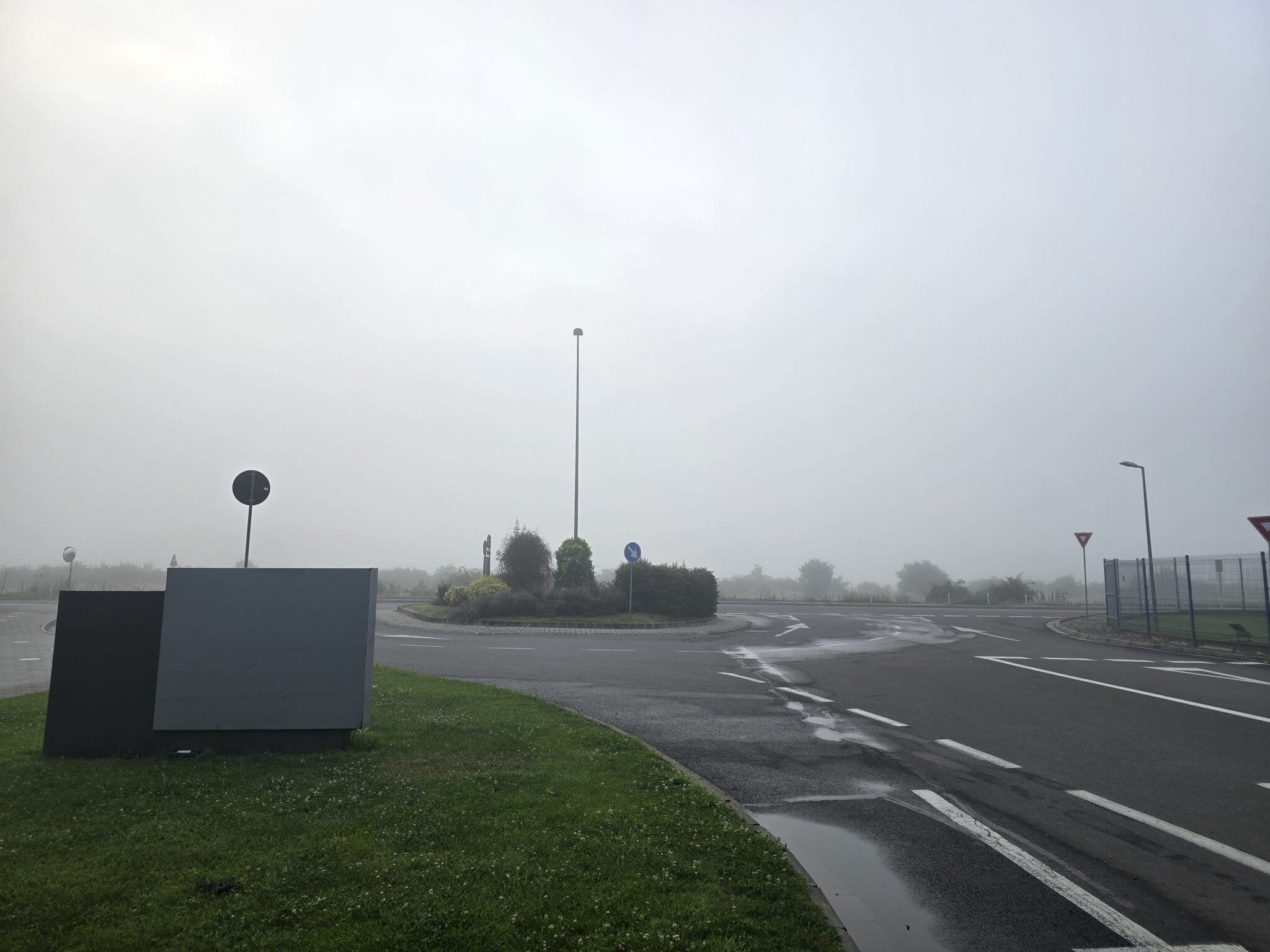 Intersecție giratoriu acoperită de ceață densă într-o dimineață ploioasă, cu vegetație verde în centru și carosabil umed