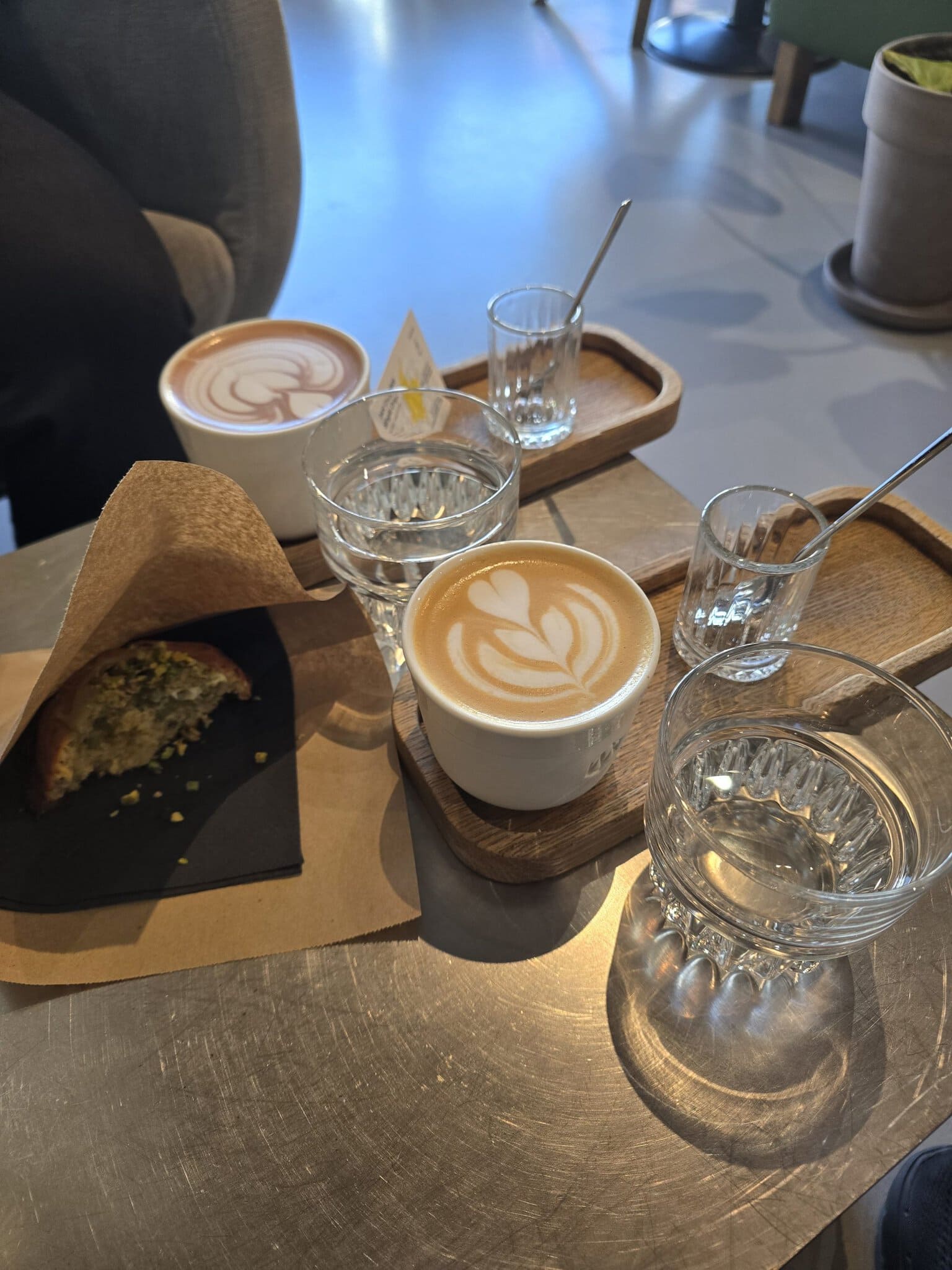 Două căni de cafea cu latte art pe o masă de metal, alături de pahare cu apă și un croissant cu fistic servit pe o tavă de lemn.