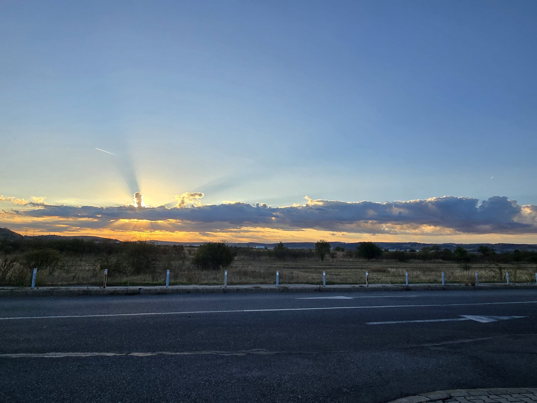 Apus de soare cu raze spectaculoase care străpung norii deasupra unui câmp arid, lângă un drum asfaltat.
