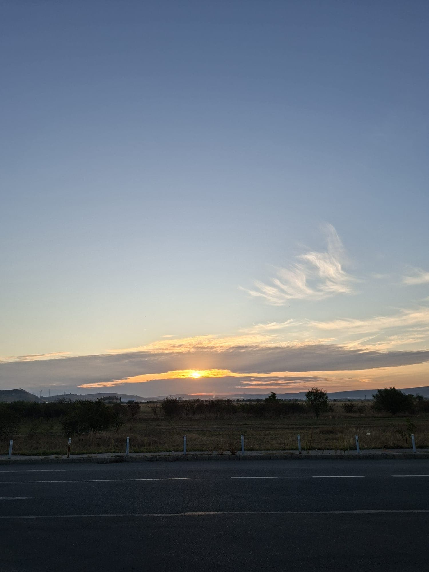Apus de soare deasupra unui câmp deschis, cu cerul colorat în nuanțe de portocaliu și albastru, văzut de lângă un drum asfaltat.