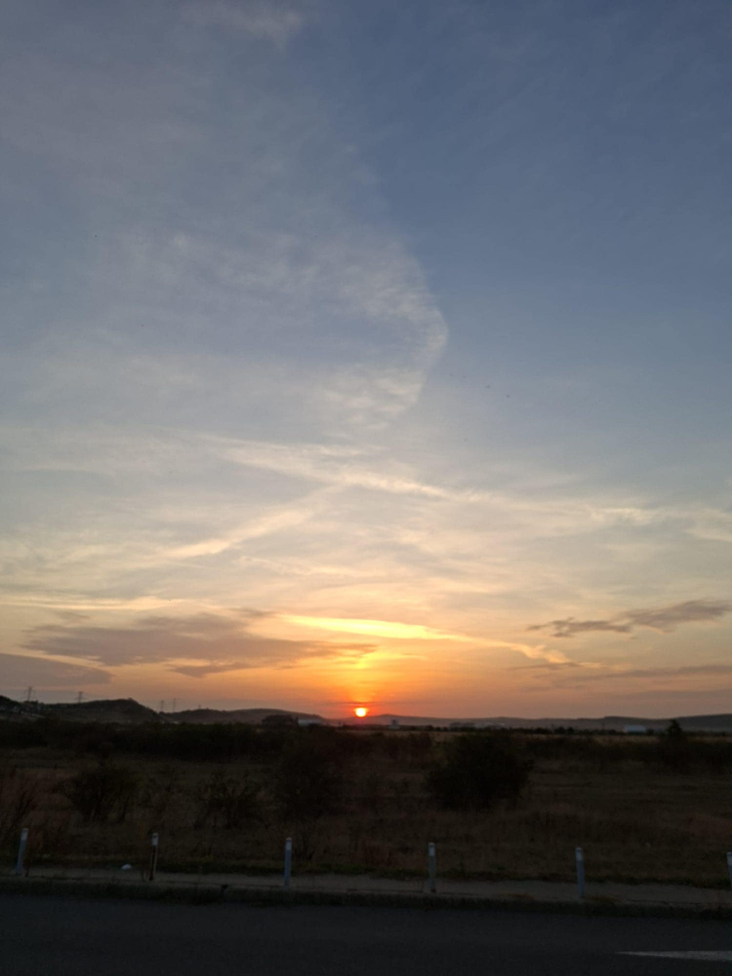 Apus de soare portocaliu deasupra unui câmp arid și a unor dealuri line, cu nori subțiri pe cerul albastru.