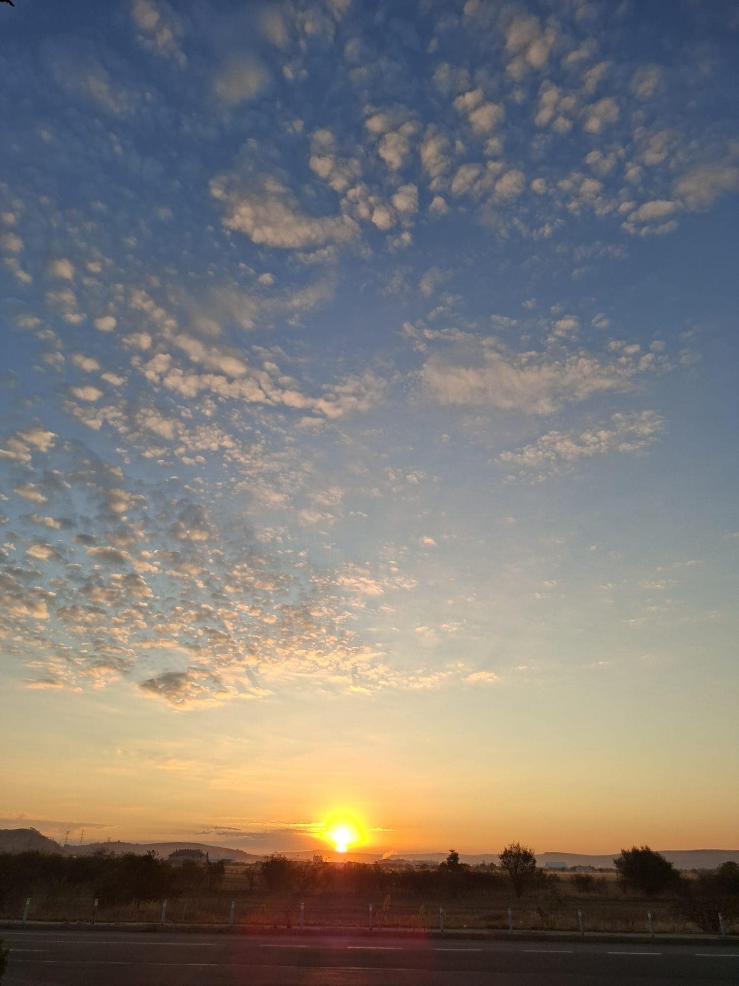 Apus de soare portocaliu intens pe orizont, cu nori altocumulus împrăștiați pe cerul albastru, deasupra unui drum și a unor copaci în silhouetă.