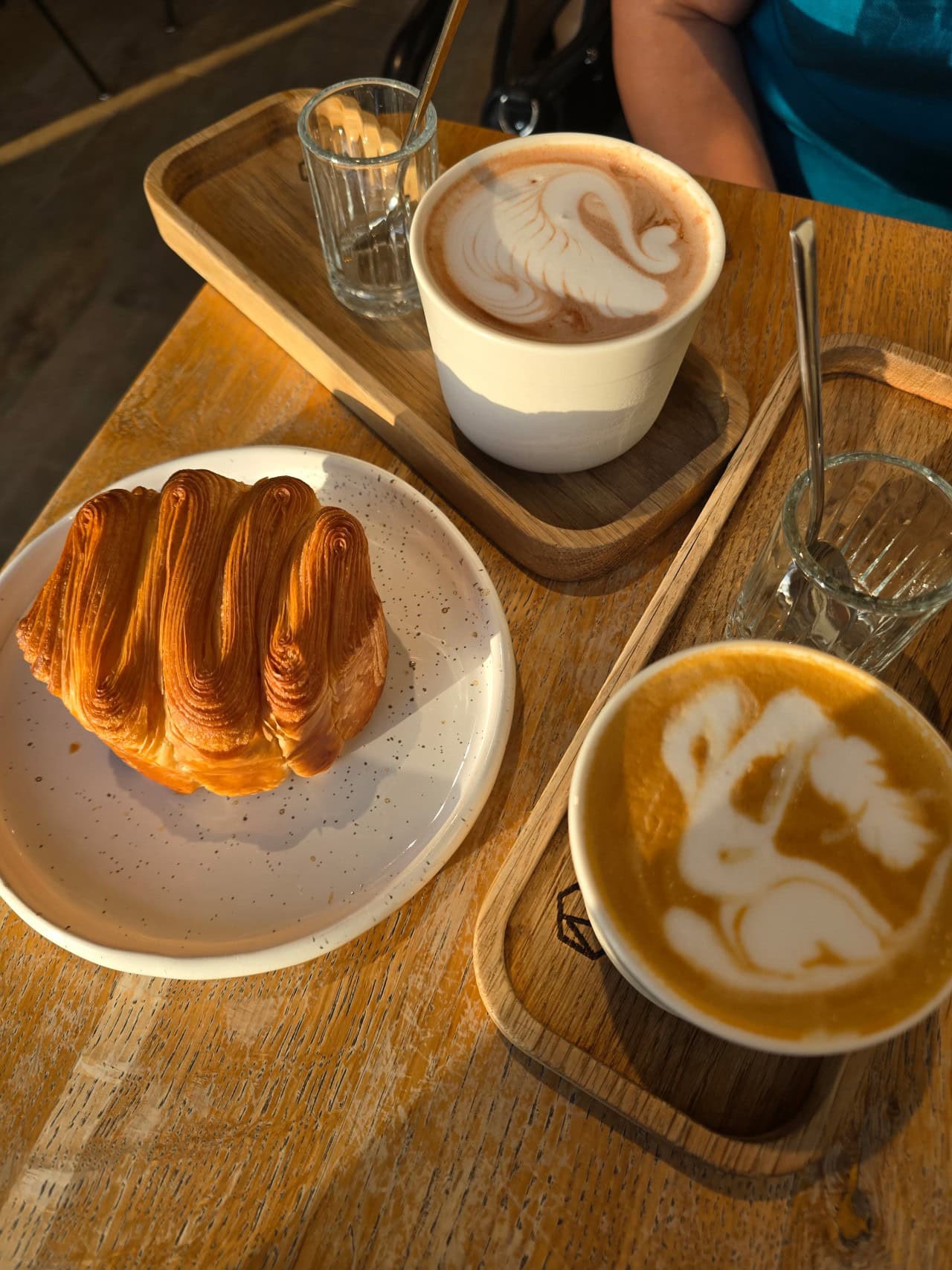 Două căni de cafea cu latte art și un croissant auriu pe o tavă de lemn, fotografiate de sus.