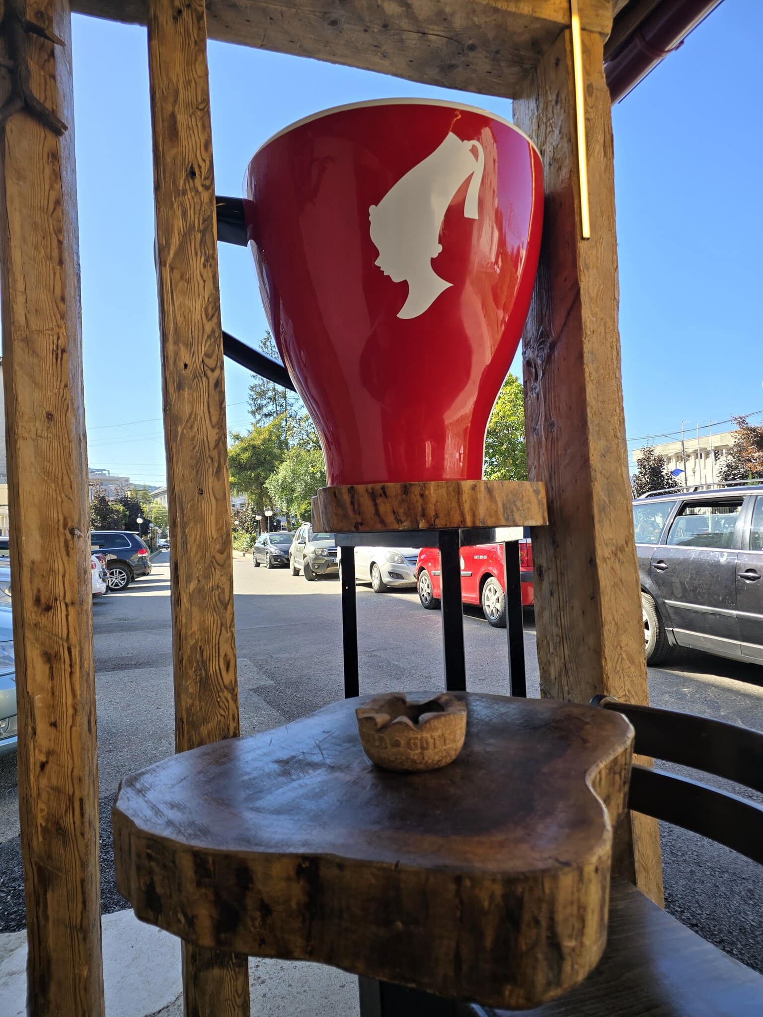 O cană gigantică roșie cu silueta mărcii Julius Meinl, expusă pe o terasă din lemn rustic în fața unei străzi.