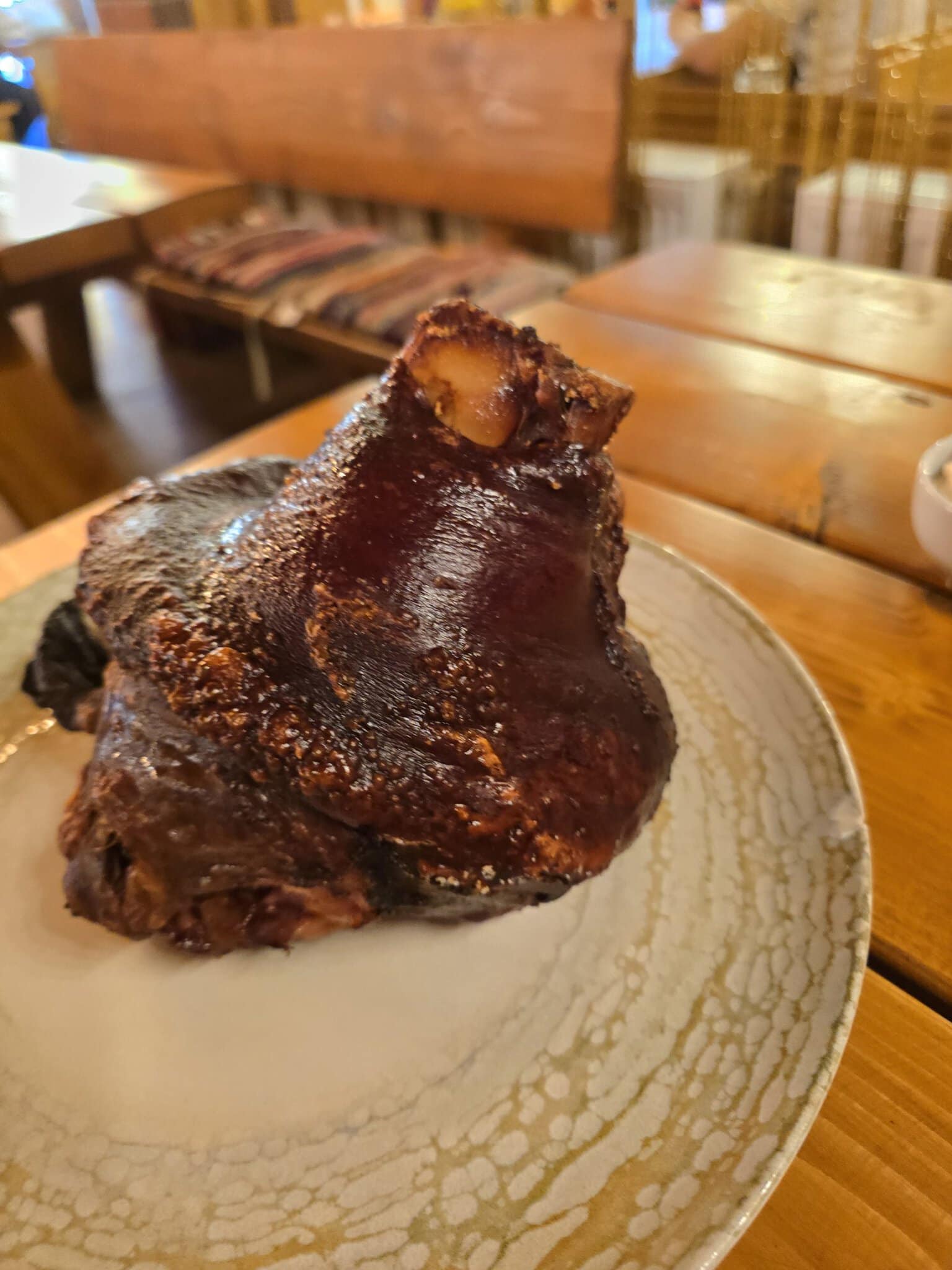 Un cotlet de porc afumat și glazurat, servit pe o farfurie ceramică albă cu model, într-un restaurant cu mobilier din lemn.