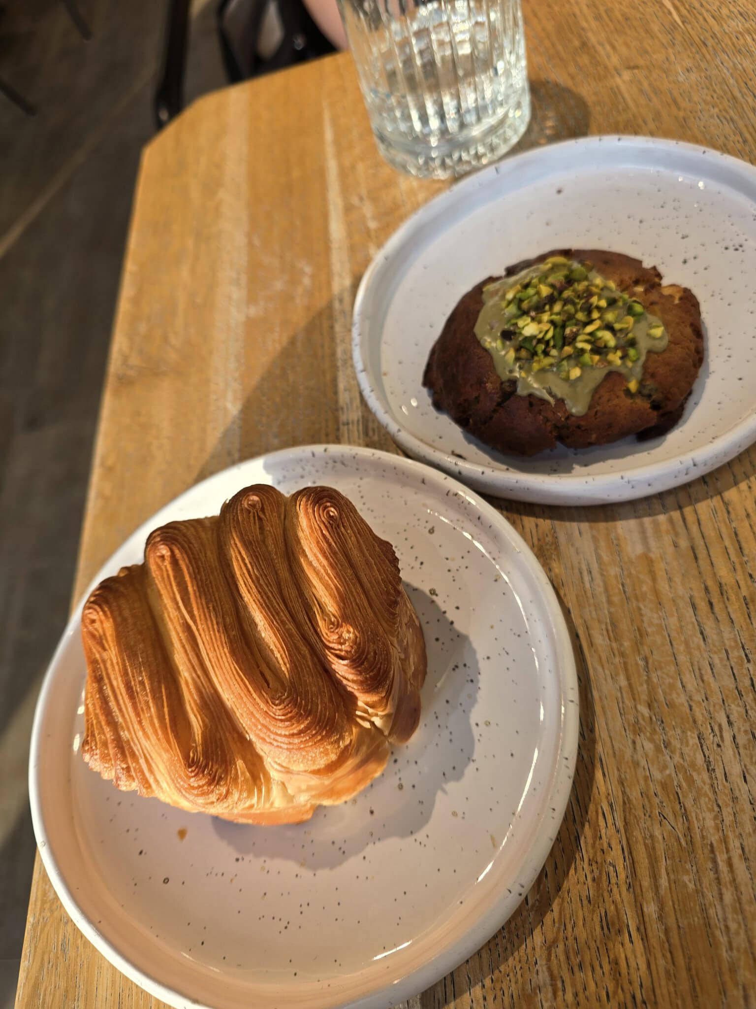 Două produse de patiserie pe farfurii albe cu puncte, un croissant pufos și un cookie cu glazură de fistic și fistic tocat, așezate pe o masă de lemn într-o cafenea.