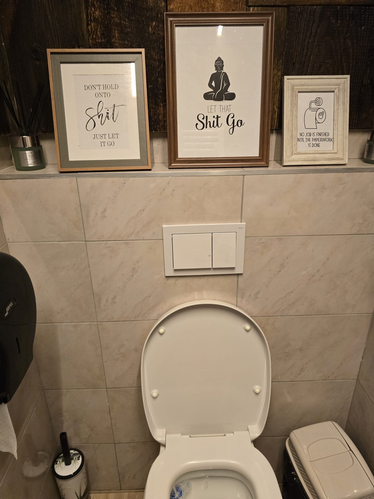 Trei tablouri înrămate cu mesaje amuzante despre toaletă, expuse pe un raft deasupra unui vas WC alb, pe un fundal de gresie marmurată și lemn închis la culoare.