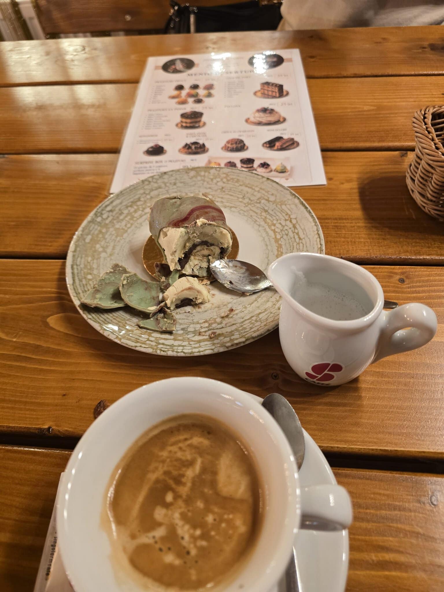 O prăjitură colorată cu cremă și glazură verde, parțial consumată, alături de o ceașcă de cafea cu lapte și un ulcior mic, pe o masă de lemn într-o cafenea.