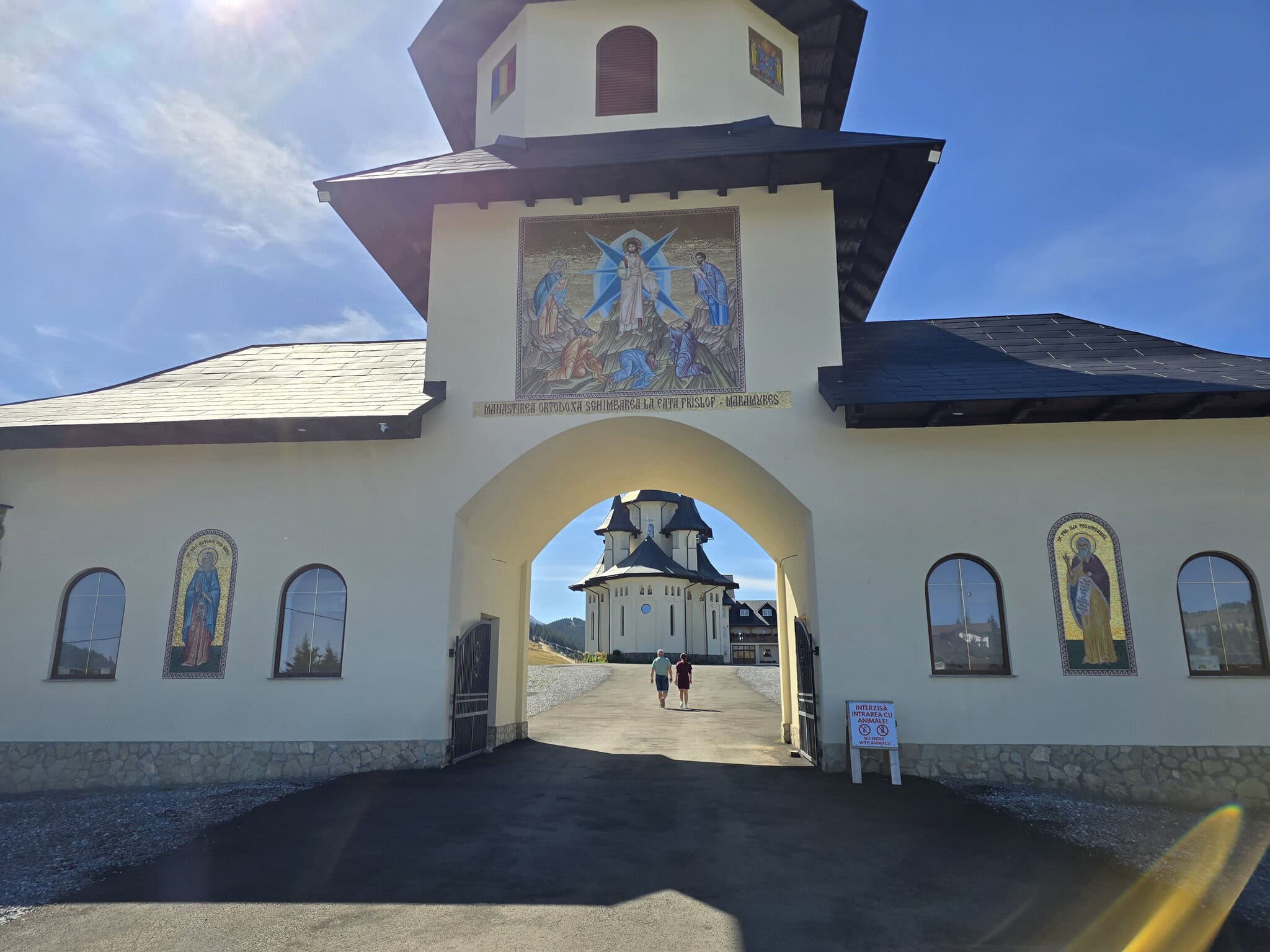 Poarta de intrare a Mănăstirii Ortodoxe Schimbarea la Față de la Prislop, Maramureș, cu mozaic religios și doi vizitatori în curte.