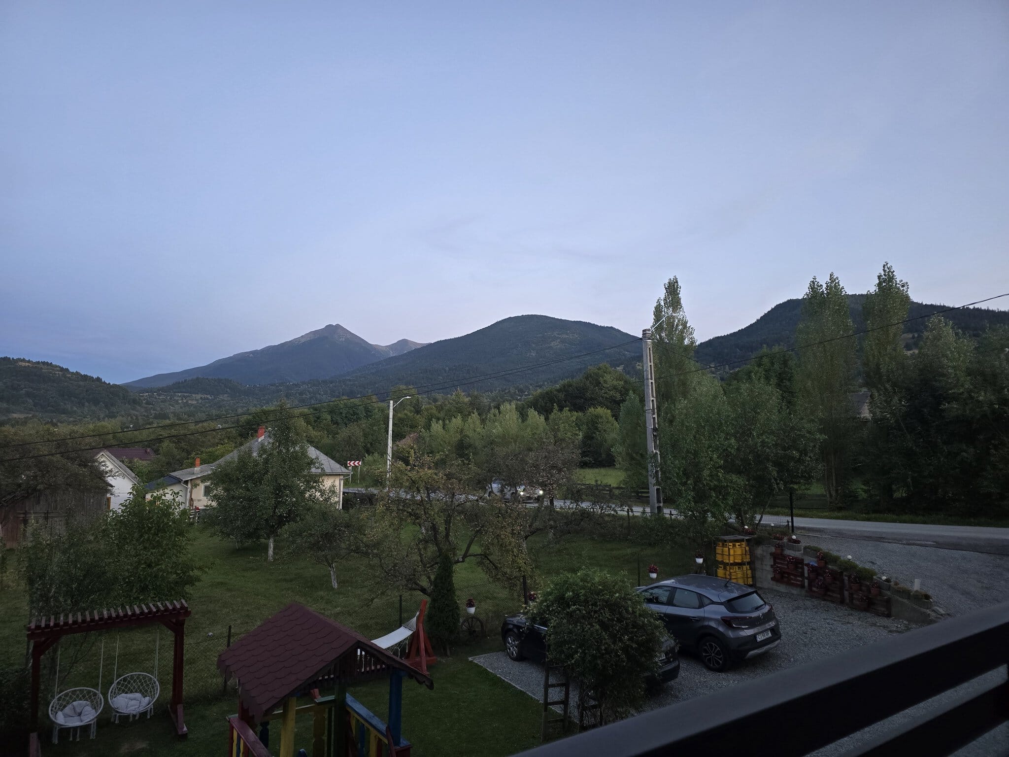 Vedere de pe balcon spre munții Carpați la amurg, cu case și arbori în prim-plan și o mașină parcată pe alee cu pietriș.