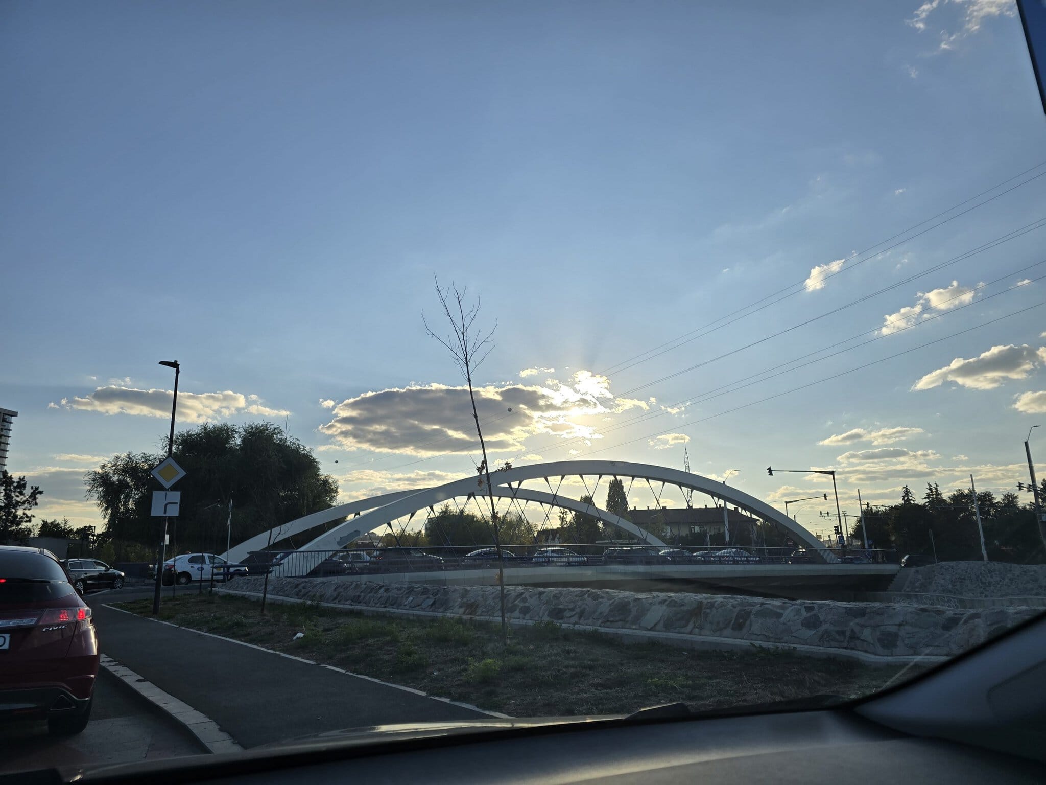 Pod arcuit alb fotografiat din mașină în lumina caldă a apusului, cu trafic și cer parțial înnorat în fundal.