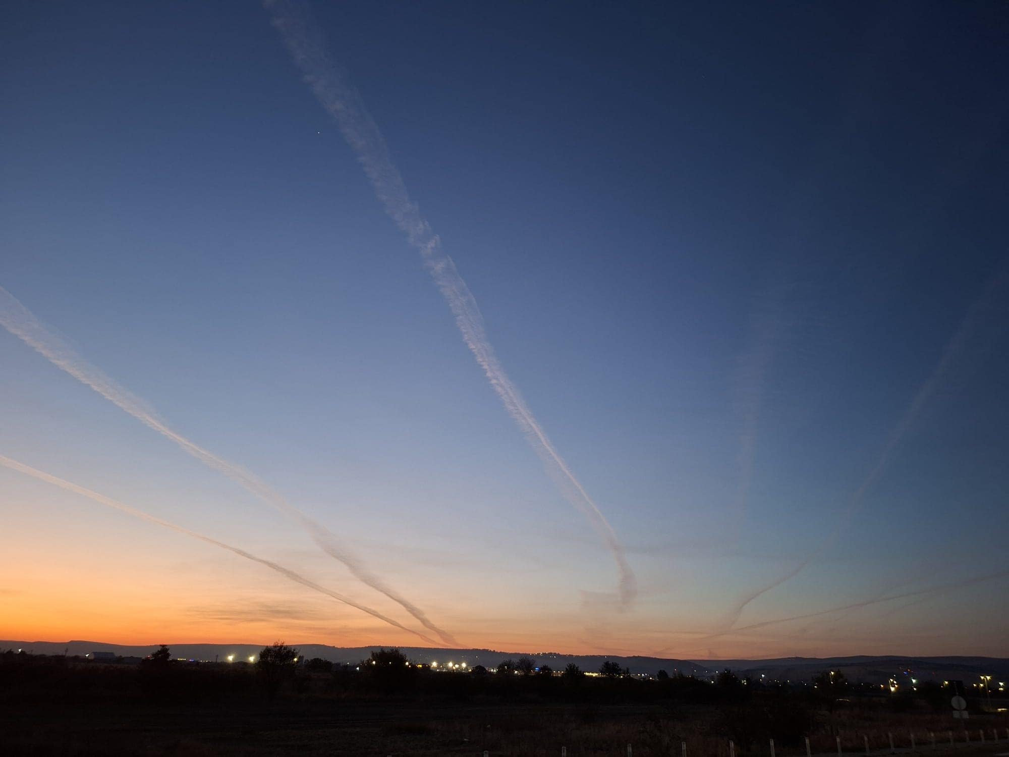 Apus de soare cu urme de condensare ale avioanelor pe cerul albastru, deasupra unui orizont luminat de luminile orașului.