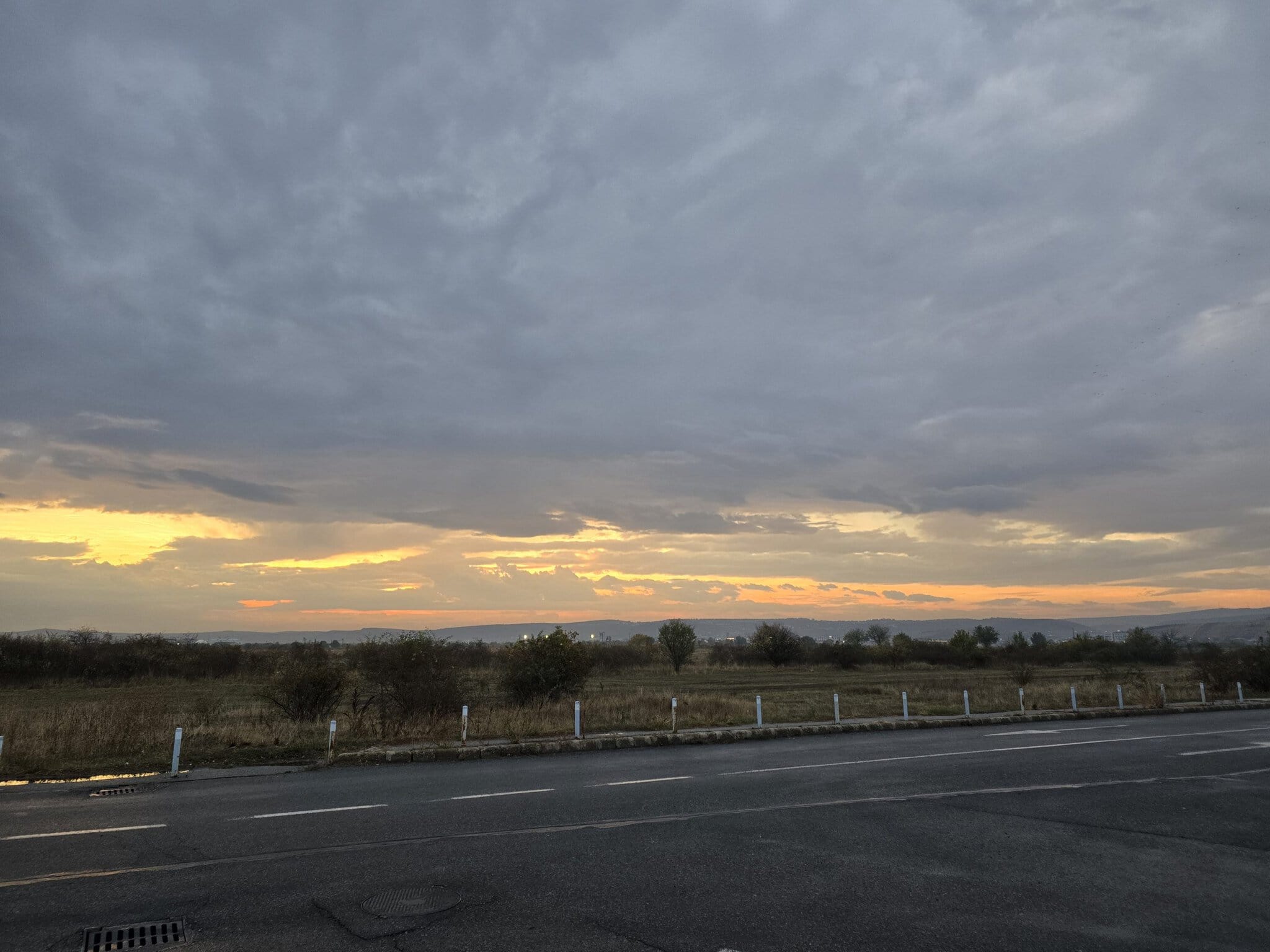 Un apus de soare cu nuanțe portocalii și galbene printre nori cenușii, deasupra unui câmp arid de toamnă, fotografiat de lângă o șosea cu marcaje rutiere albe.