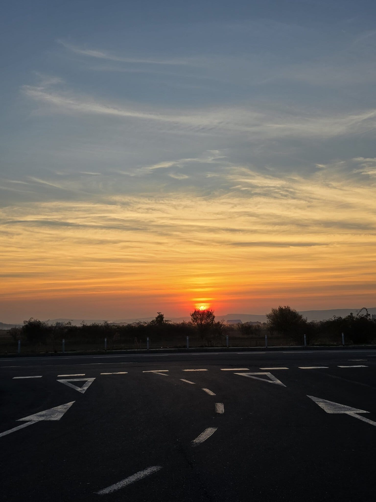 Apus de soare spectaculos văzut de pe autostradă, cu cerul colorat în nuanțe de portocaliu și roșu deasupra unui șir de copaci și dealuri la orizont.