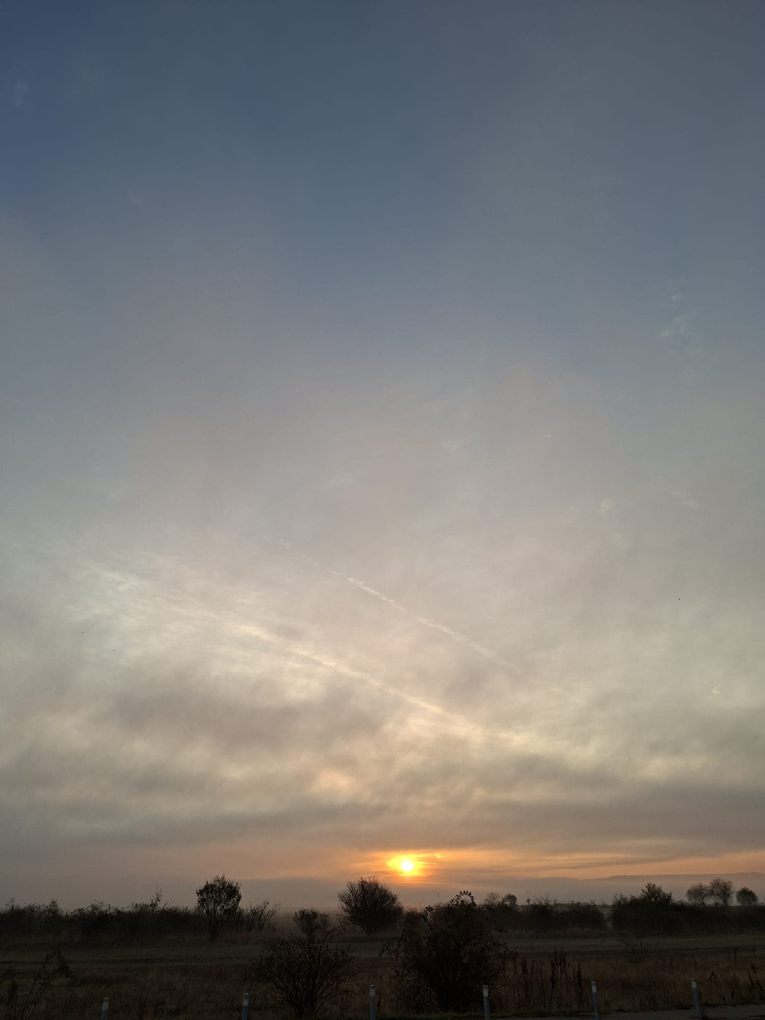 Soarele apune la orizont printre nori, deasupra unei câmpii cu copaci desfrunziți.