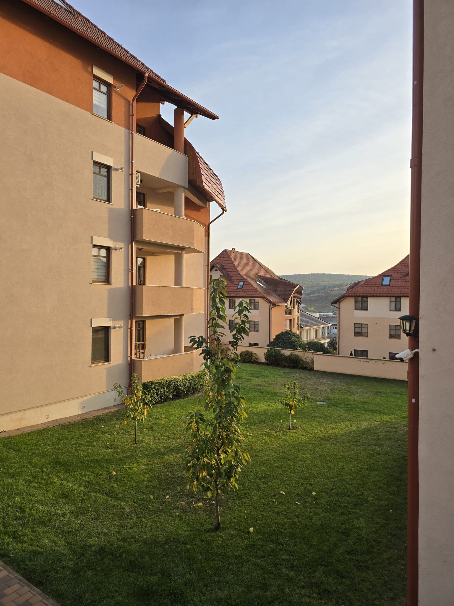 Vedere din curtea unui complex rezidențial cu blocuri bej și acoperiș roșu, gazon verde și dealuri în fundal la apus.