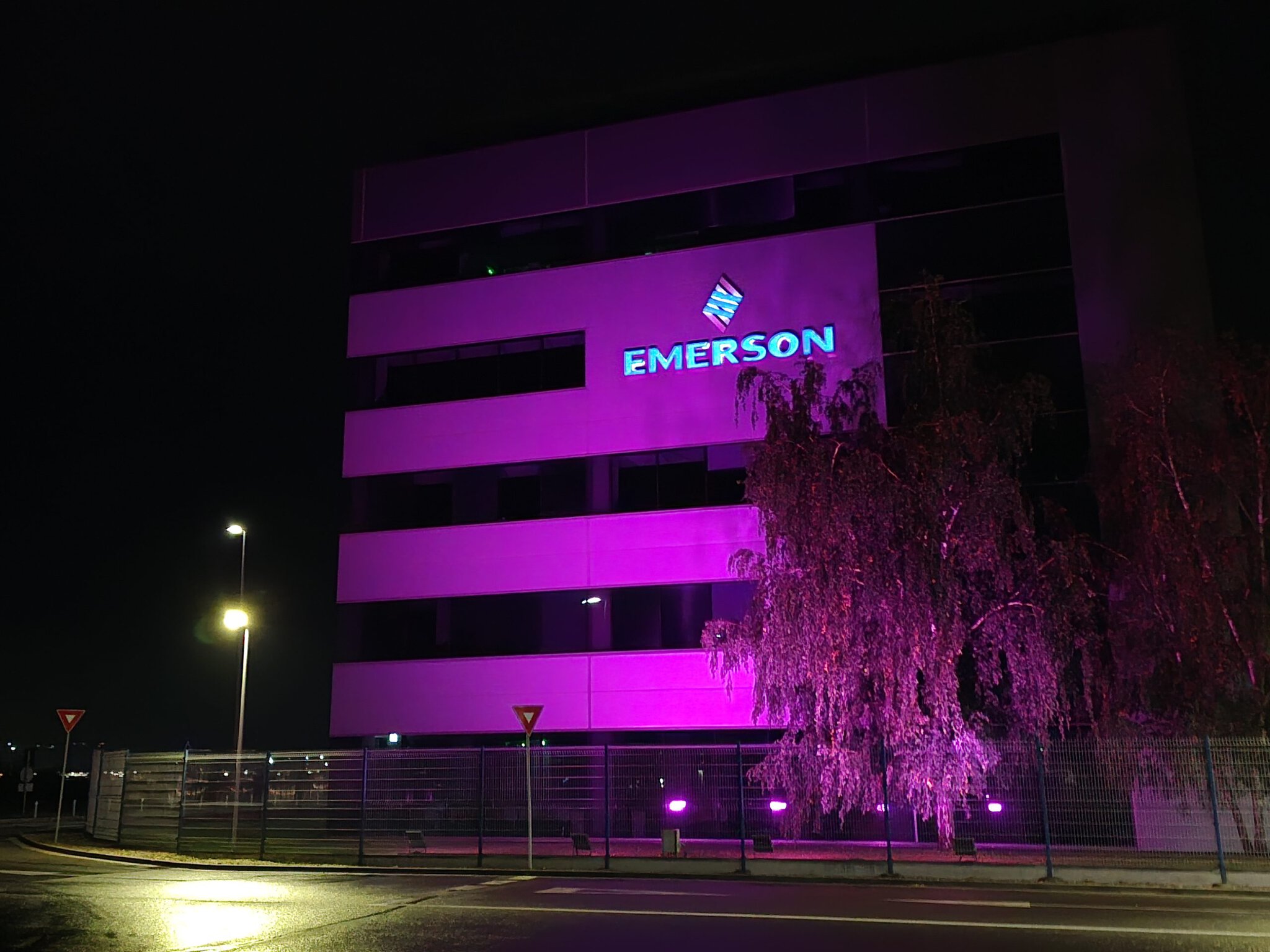 Clădirea Emerson iluminată în mov și magenta pe timp de noapte, cu un copac decorativ în față și logo luminos albastru.