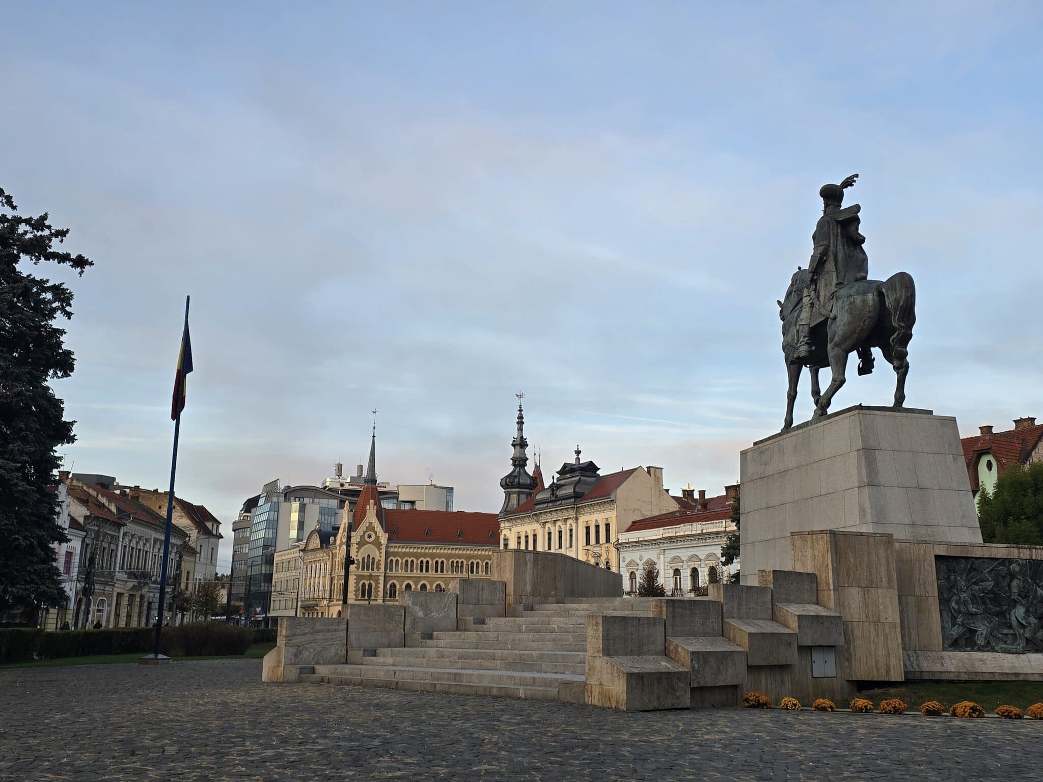 Statuia ecvestră din Piața Avram Iancu din Cluj-Napoca fotografiată în lumina dimineții, cu clădiri istorice și drapelul României în fundal.